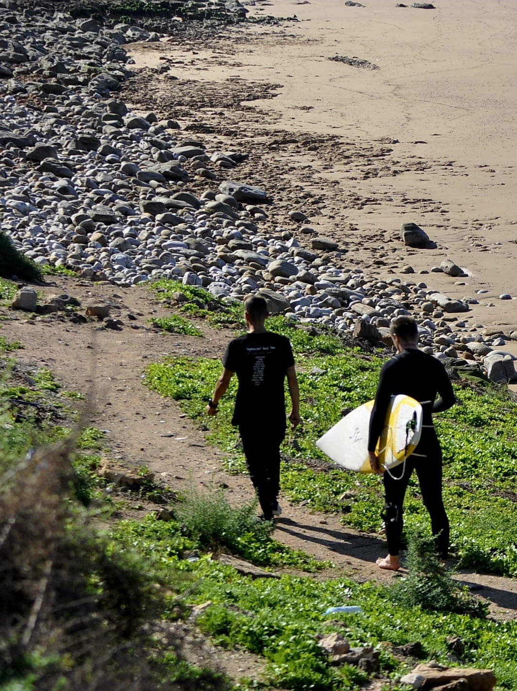 Two surfers walking down a coastal path toward K12